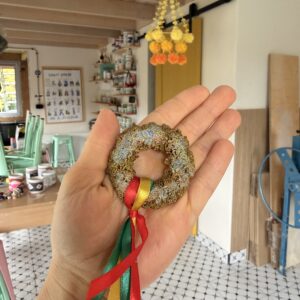 Hand holding a beaded circular ornament tied with red, green, and yellow ribbons in a craft studio.