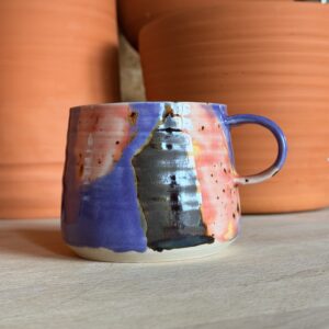 Colorful glazed ceramic mug on a light wooden surface with large terracotta pots in the background
