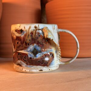 Close-up of a chipped ceramic mug with multicolored glaze on a wooden surface, with bright orange chairs blurred in the background