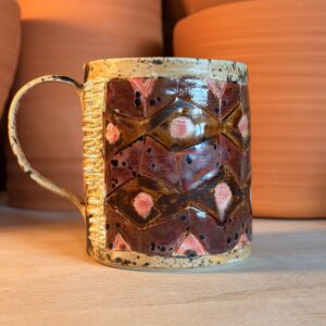 Ceramic mug with brown geometric pattern and a textured pale handle on a light wooden surface. Background shows orange pottery jars.