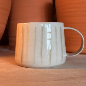 A light beige ceramic mug sits on a wooden surface with large orange objects in the background.